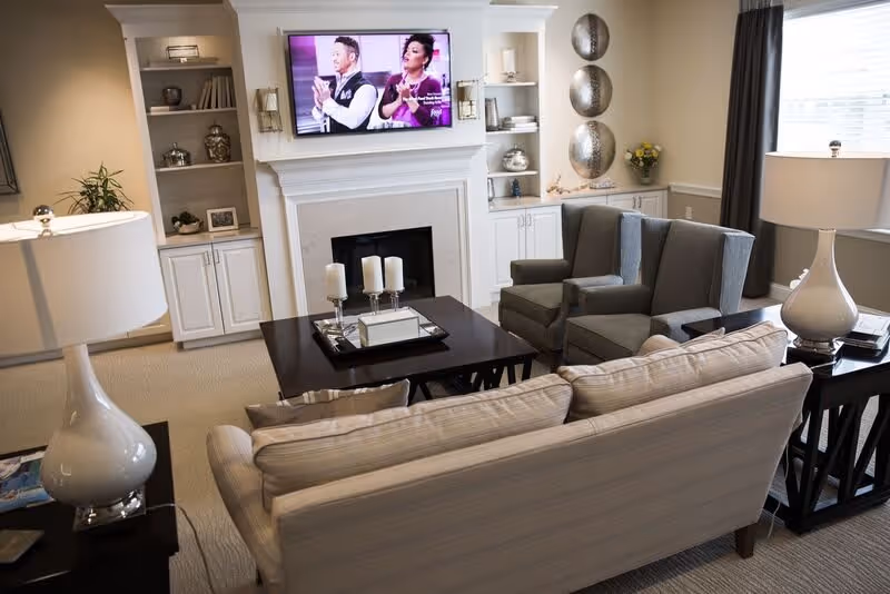 A cozy living room with a beige sofa and two gray armchairs arranged around a dark wooden coffee table with candles and a tray. A flat-screen TV is mounted above a white fireplace mantel. Built-in shelves flank the fireplace, decorated with books, vases, and other decorative items. Two table lamps with white shades are on side tables next to the sofa and armchairs. A window with dark curtains lets in natural light.