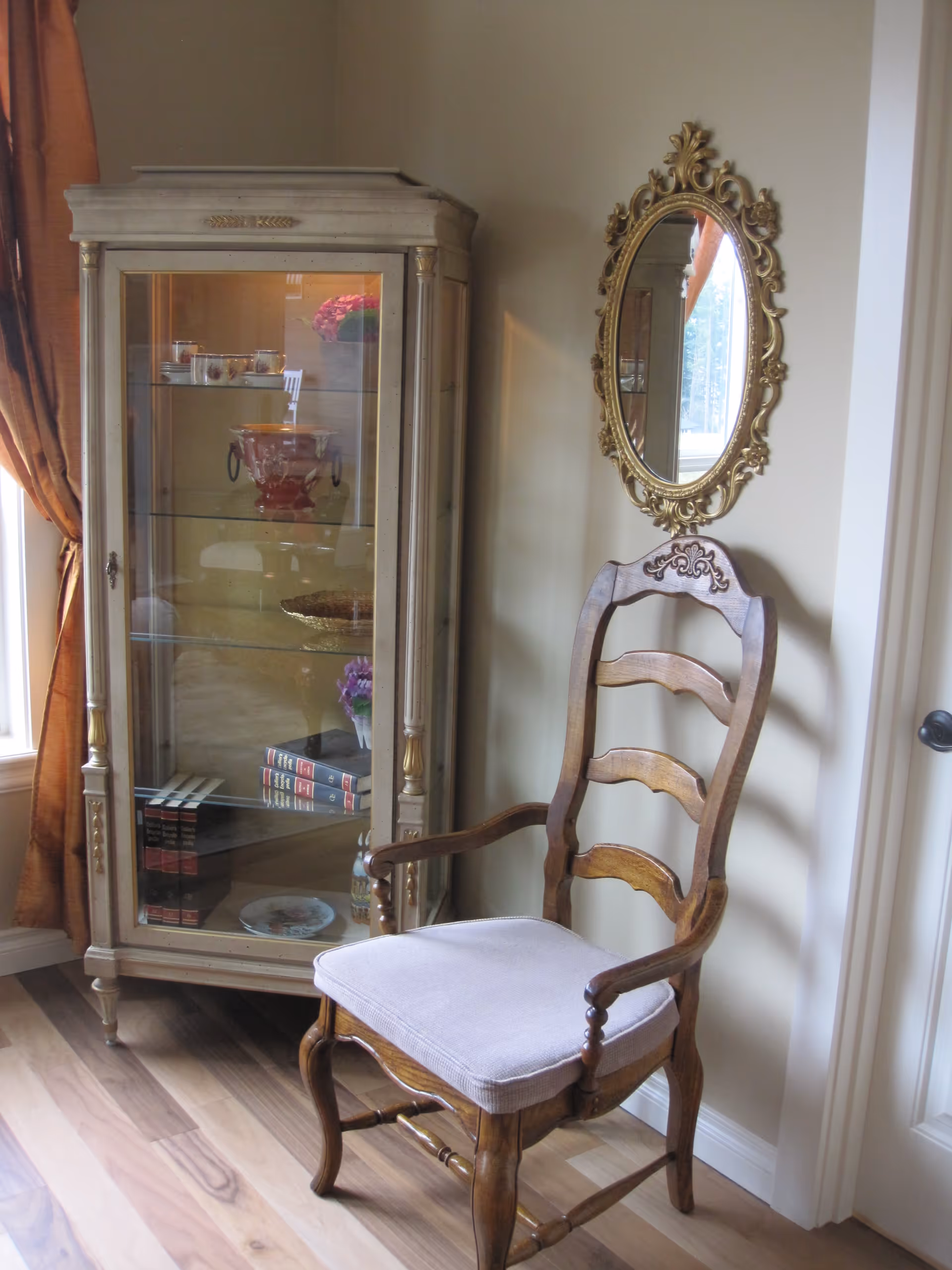 A corner of a room featuring a wooden chair with a cushioned seat, an ornate gold-framed oval mirror on the wall, and a glass display cabinet containing decorative items and books. The room has light-colored walls and wood flooring, with a window partially covered by a brown curtain.