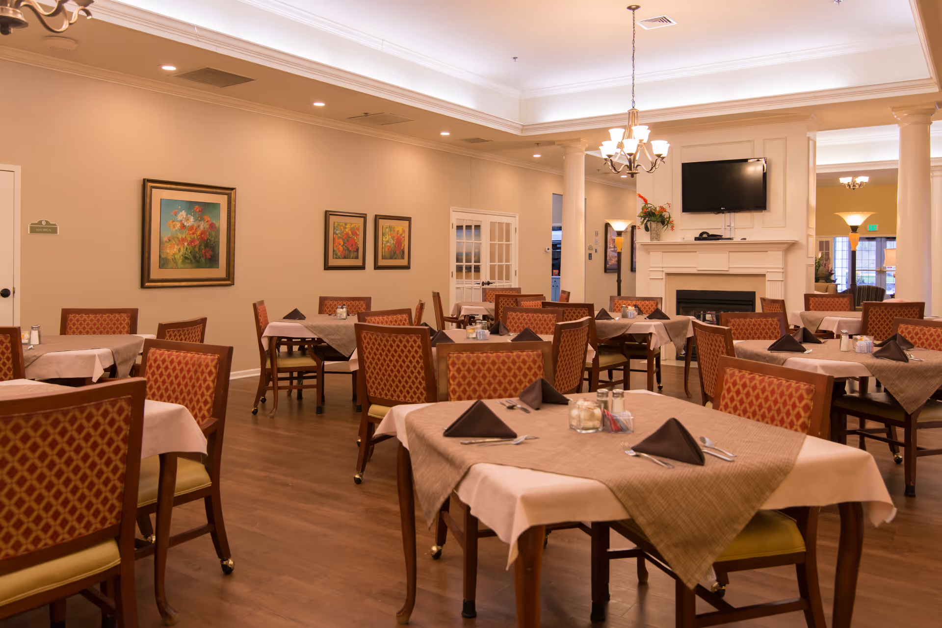 Spacious dining room with multiple set tables and chairs, a fireplace with wall-mounted TV, and framed artwork on the walls.