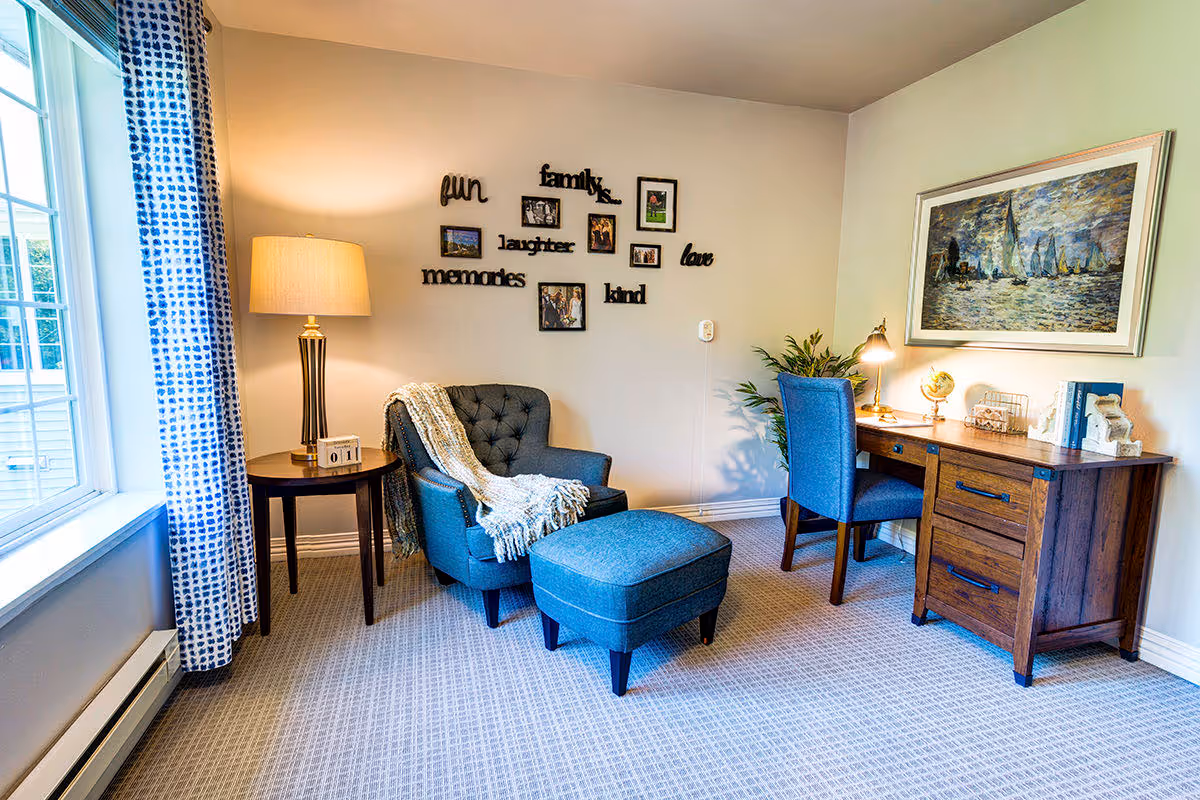 A cozy interior room with a blue upholstered armchair and matching ottoman next to a wooden side table with a lamp. On the wall above the chair are decorative words and framed photos. To the right is a wooden desk with a blue chair, a desk lamp, books, and decorative items. A large window with blue patterned curtains lets in natural light.