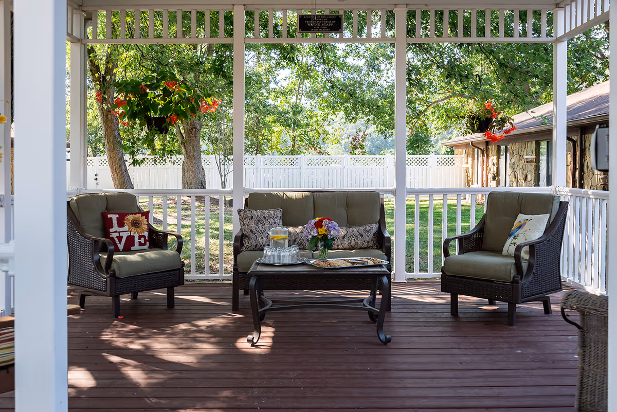 Covered outdoor patio with wicker seating and a coffee table on a wooden deck overlooking a fenced yard.