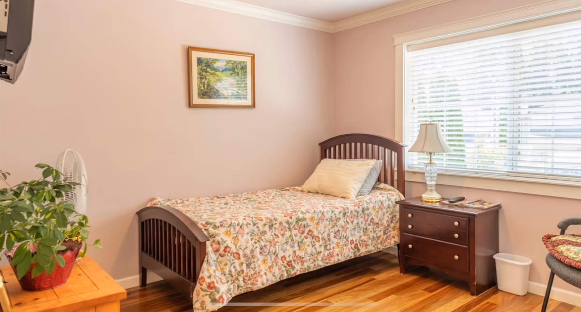 A cozy bedroom with a single wooden bed covered with a floral bedspread and two pillows. Next to the bed is a dark wooden nightstand with a decorative lamp, a remote control, and some magazines. A large window with white blinds lets in natural light. There is a framed landscape painting on the wall above the bed, a small wooden table with a potted plant and a fan, and a chair with a cushion in the corner.