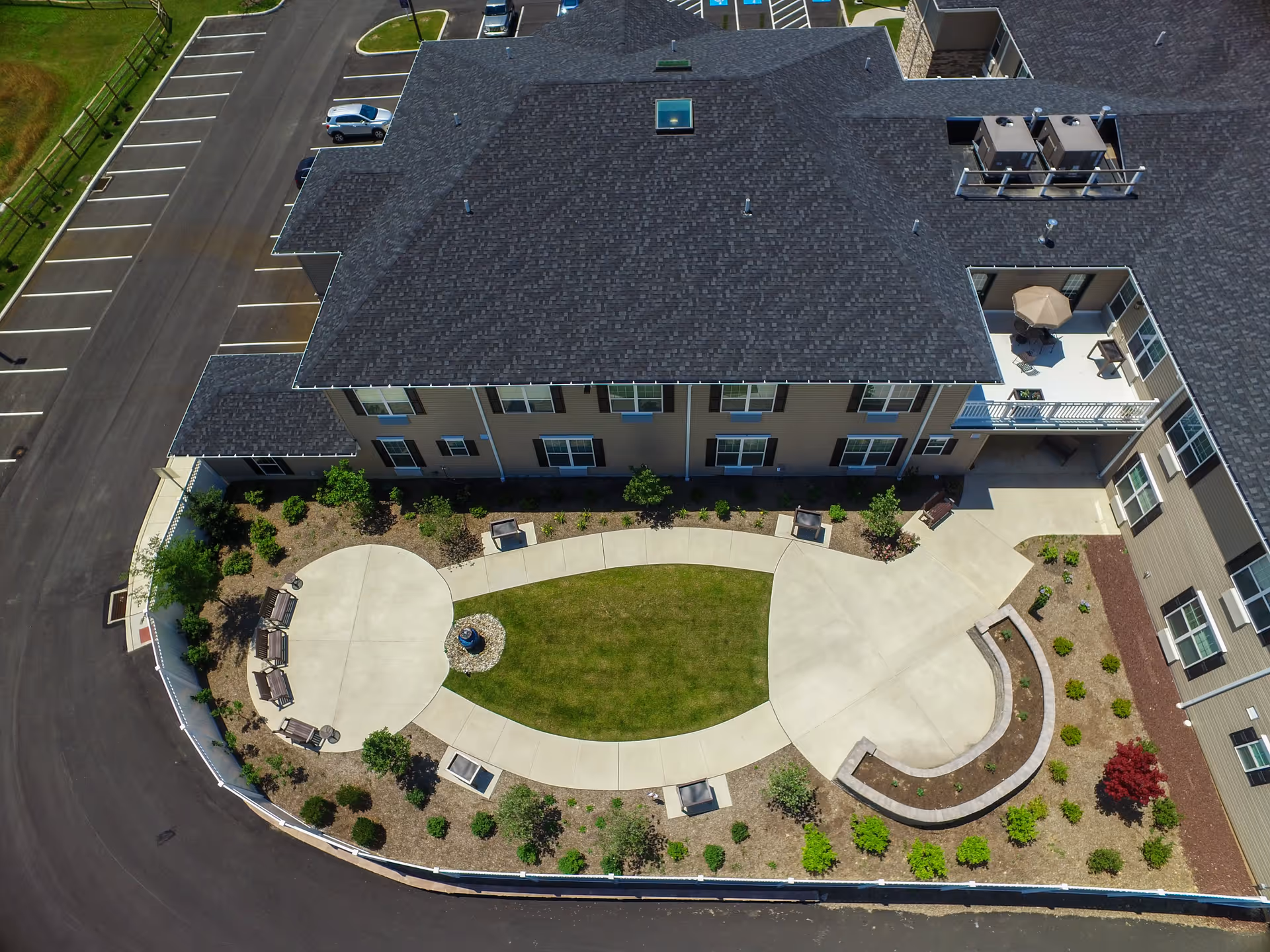 Aerial view of an outdoor courtyard area at Heather Glen Senior Living, featuring a curved concrete walkway surrounding a grass lawn, benches, landscaped plants, and a patio with an umbrella table attached to the building.