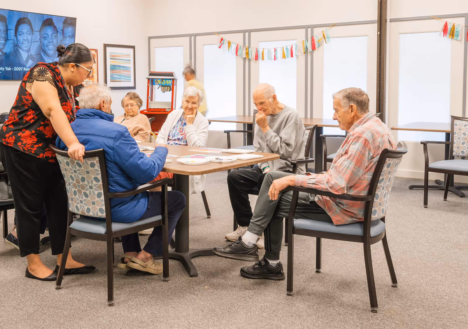 A group of elderly people sitting around a table in a well-lit room, engaging in an activity with a caregiver standing beside them. The room has large windows with white blinds, colorful garlands hanging above, and a popcorn machine in the background.