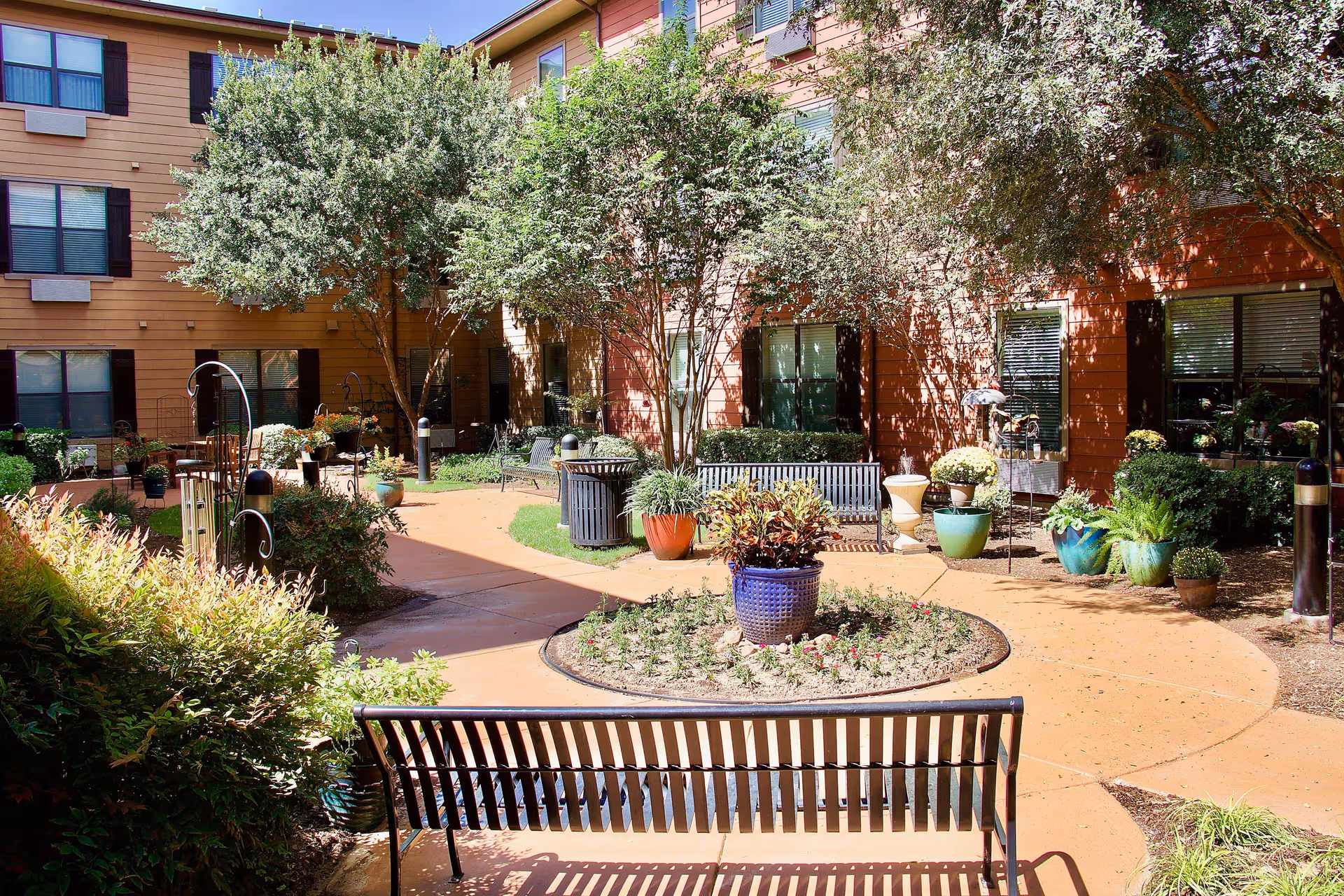 Outdoor courtyard area of a senior living facility with benches, potted plants, trees, and a circular flower bed surrounded by a paved walkway. The building exterior is visible with windows and brown siding.