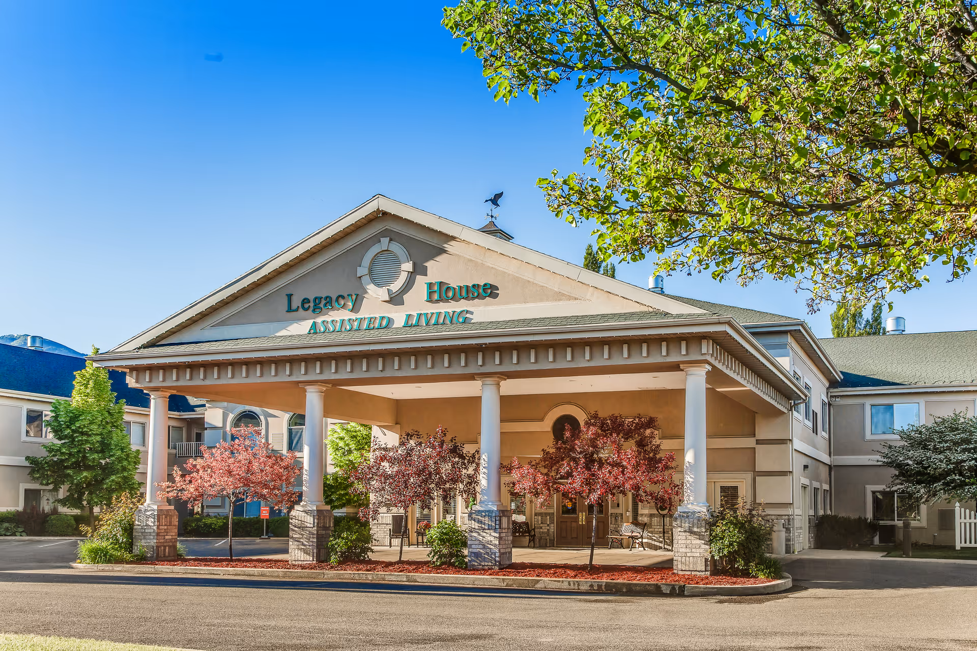Front exterior view of Legacy House Assisted Living facility with a covered entrance supported by white columns, surrounded by trees and landscaping under a clear blue sky.