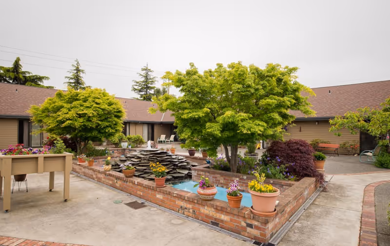 Outdoor courtyard area with a brick water feature surrounded by potted plants and flowers. There are green trees and shrubs around the water feature. The courtyard is paved with concrete and has a few benches and chairs near the building walls. The building has a brown roof and beige siding.