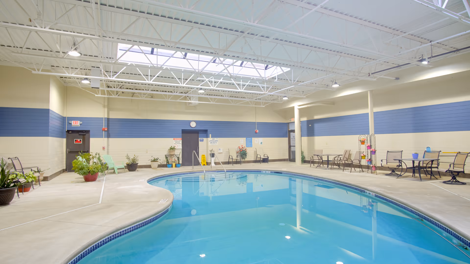 Indoor swimming pool area with clear blue water, surrounded by a concrete deck with several chairs and tables. The walls are painted beige with a blue stripe, and the ceiling has exposed white beams with skylights allowing natural light. There are potted plants around the pool area and safety signs on the walls.