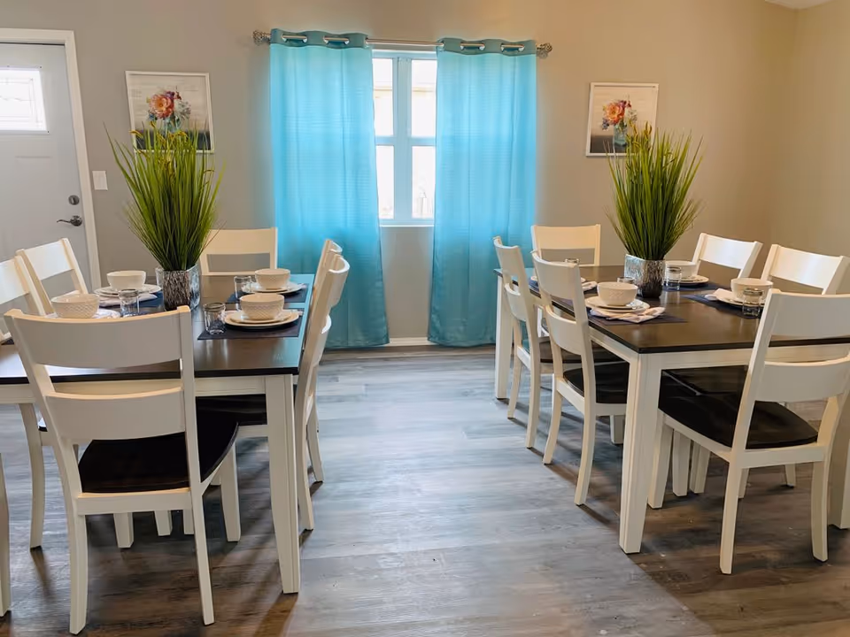 Bright dining room with two dark-top tables, white chairs, place settings and turquoise curtains at a central window.