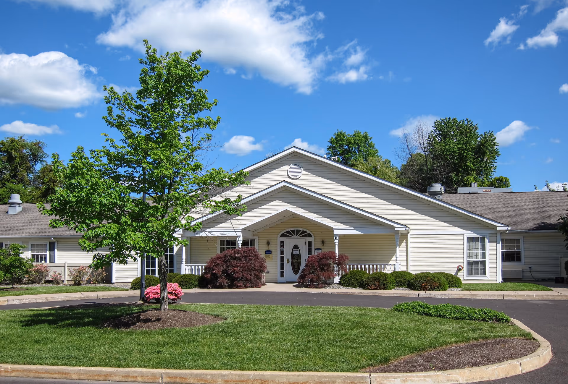 Front exterior view of a single-story building with beige siding and a peaked roof under a blue sky with scattered clouds. There is a small covered porch at the entrance with white railings, surrounded by green bushes and a tree with fresh green leaves in the foreground. The driveway curves around a grassy area with mulch beds.