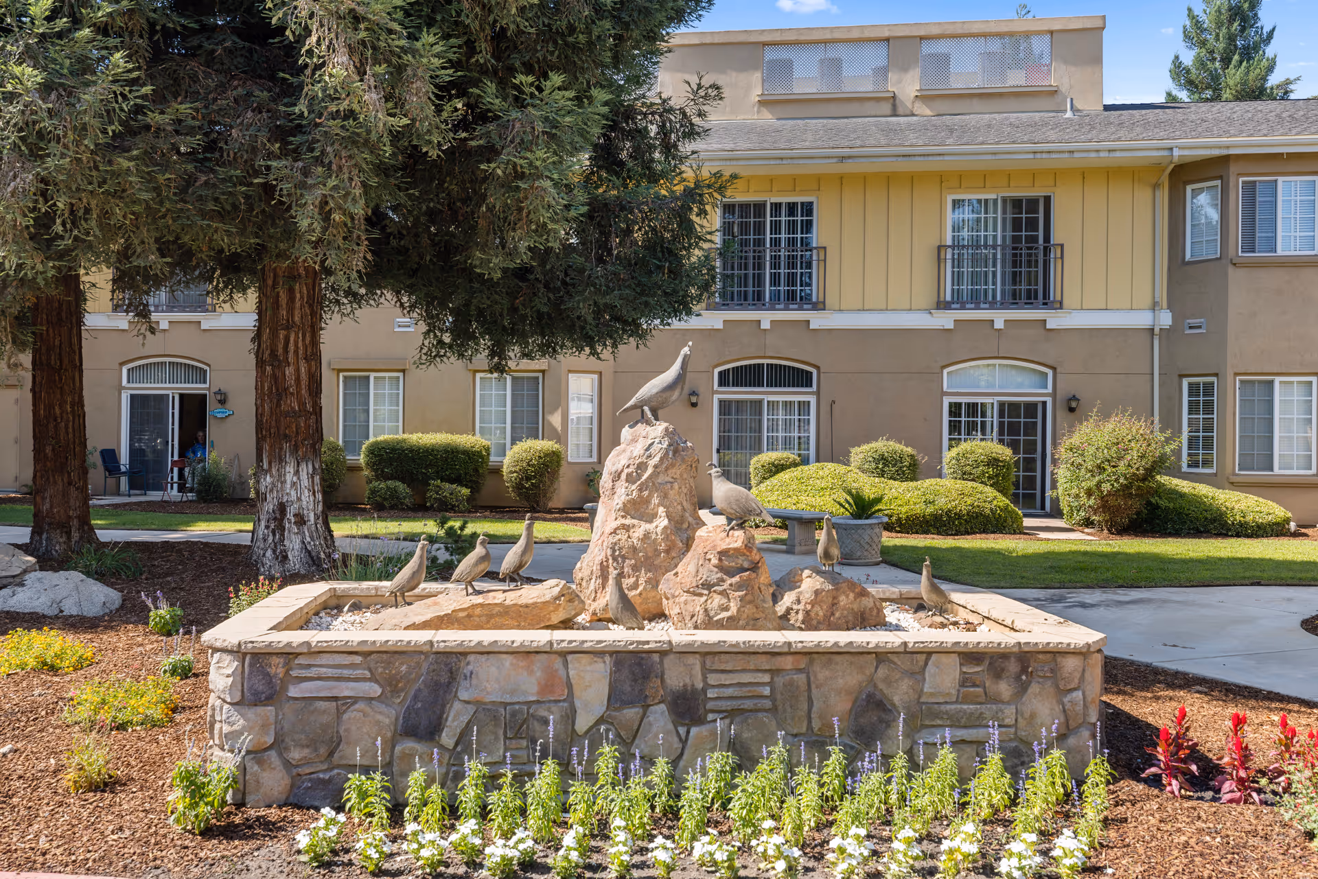 Outdoor garden area at Quail Park on Cypress featuring a stone planter with rock sculptures of birds, surrounded by flowers and greenery, with a beige multi-story building and trees in the background.