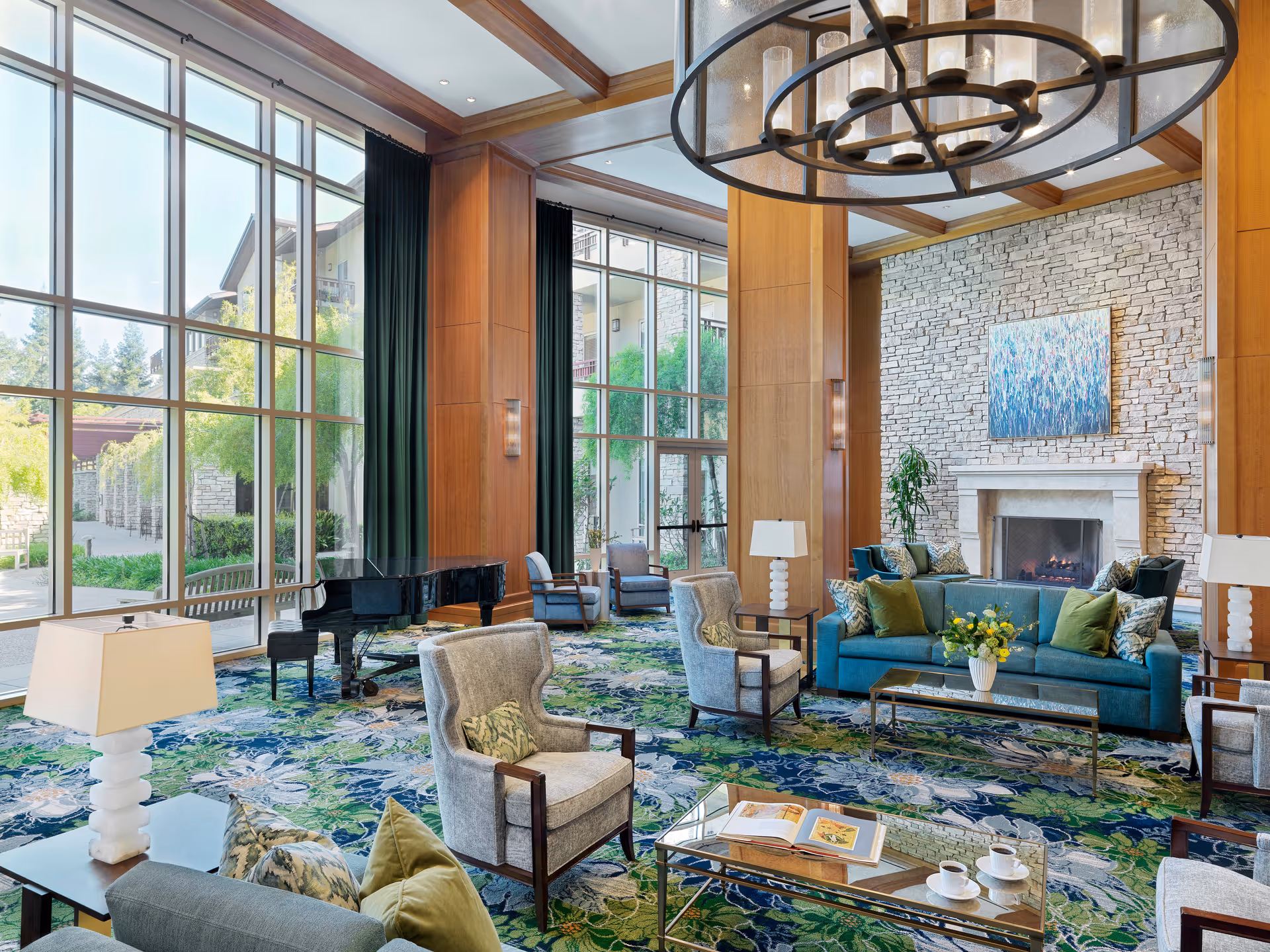 A spacious and elegant living room with large floor-to-ceiling windows allowing natural light to fill the space. The room features a blue sofa with green and patterned cushions, several armchairs, a glass coffee table with an open book and two cups, a grand piano, a stone fireplace with a colorful abstract painting above it, and decorative lamps. The carpet has a floral pattern in shades of blue and green, and wooden beams and panels add warmth to the room.