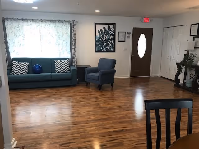 A spacious living room area with wooden flooring, featuring a teal sofa with two chevron-patterned pillows and a blue decorative ball, a single blue armchair, a small side table, a framed botanical artwork on the wall, a brown door with an oval window, white closets, and a dark wooden console table with plants and decorations.