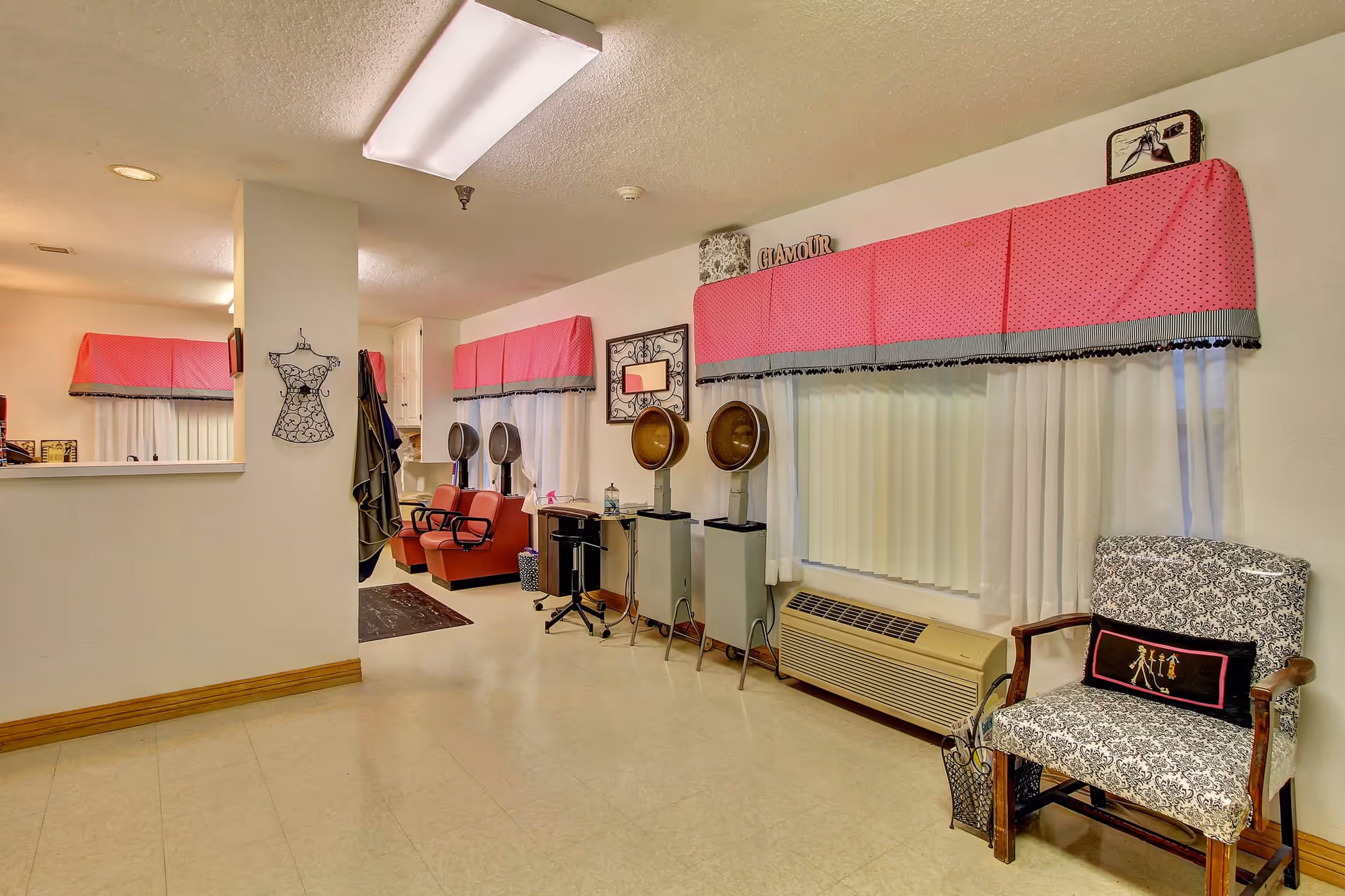Interior view of a hair salon area in a senior living facility with two vintage hair dryers, red salon chairs, a patterned armchair with a decorative pillow, pink valances over the windows, and a wall decoration shaped like a dress.