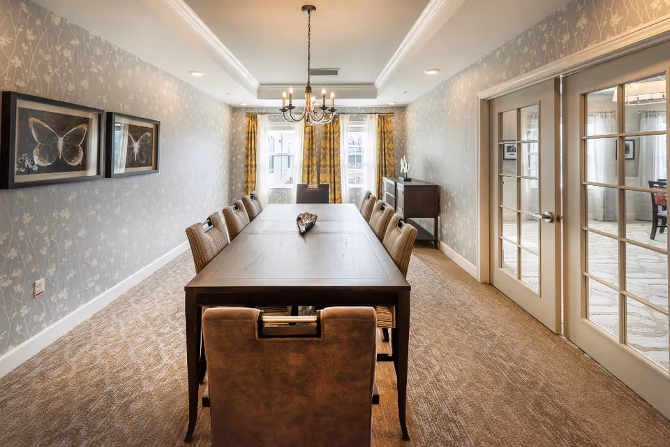 Long formal dining room with a dark wood table and upholstered chairs under a chandelier, patterned wallpaper, and French doors.