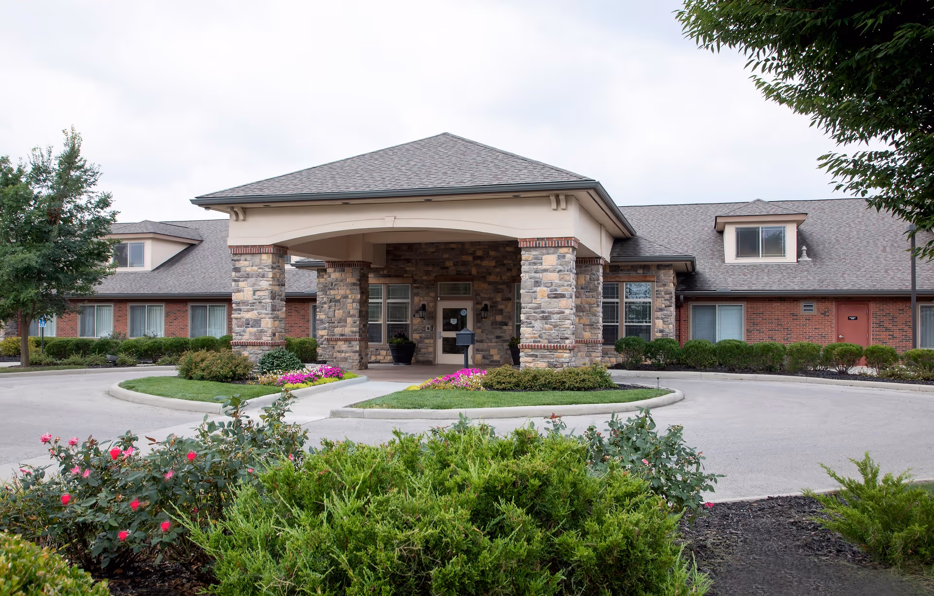 Front exterior view of a single-story senior living facility building with a covered entrance supported by stone pillars. The building has a brick facade with multiple windows and a gray shingled roof. There are landscaped bushes and flowering plants in the foreground and a circular driveway leading to the entrance.