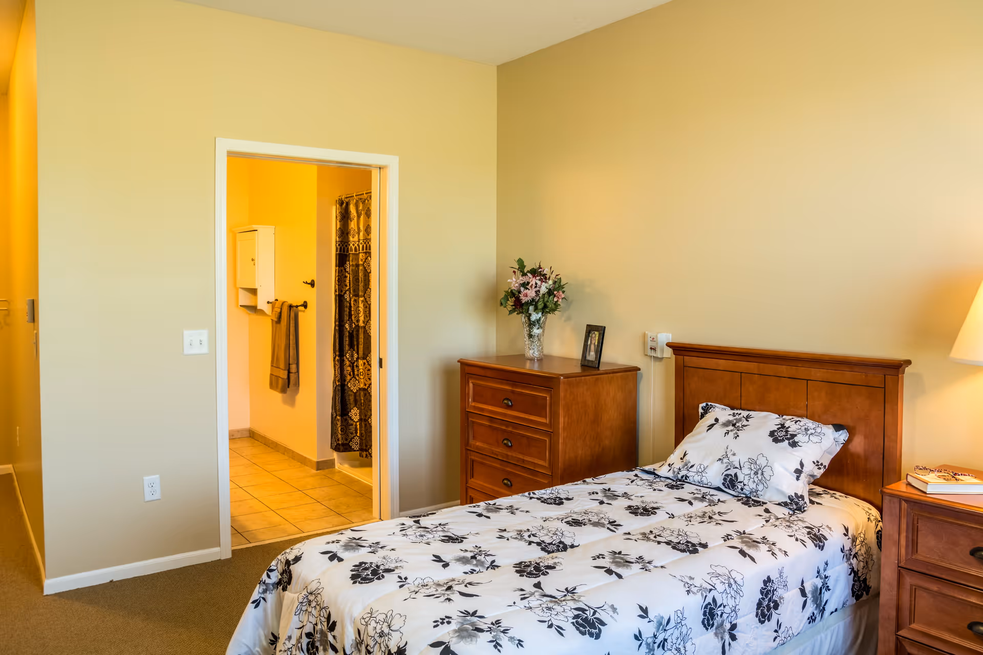 A cozy bedroom with a single bed featuring a floral patterned bedspread and pillow. Next to the bed is a wooden nightstand with a lamp and a book. There is a wooden dresser with a vase of flowers and a framed photo on top. An open door reveals a bathroom with tiled floor, a towel hanging on the wall, and a shower curtain with a decorative pattern.