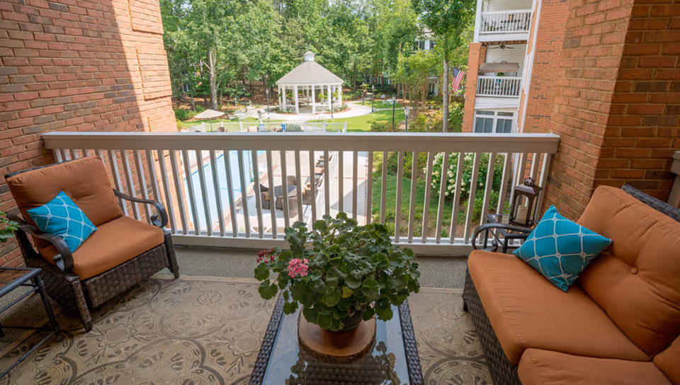 A cozy outdoor balcony with two cushioned wicker chairs and a matching loveseat, each with a blue decorative pillow. A glass-top coffee table with a potted plant sits in the center on a patterned outdoor rug. The balcony overlooks a courtyard with a white gazebo, green lawn, trees, and a swimming pool area with lounge chairs.