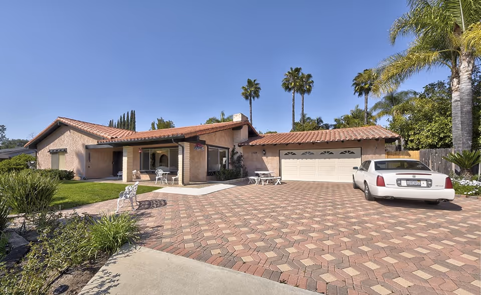 Single-story house with a tiled roof, a large paved driveway, a white car parked on the right side, and palm trees in the background under a clear blue sky.