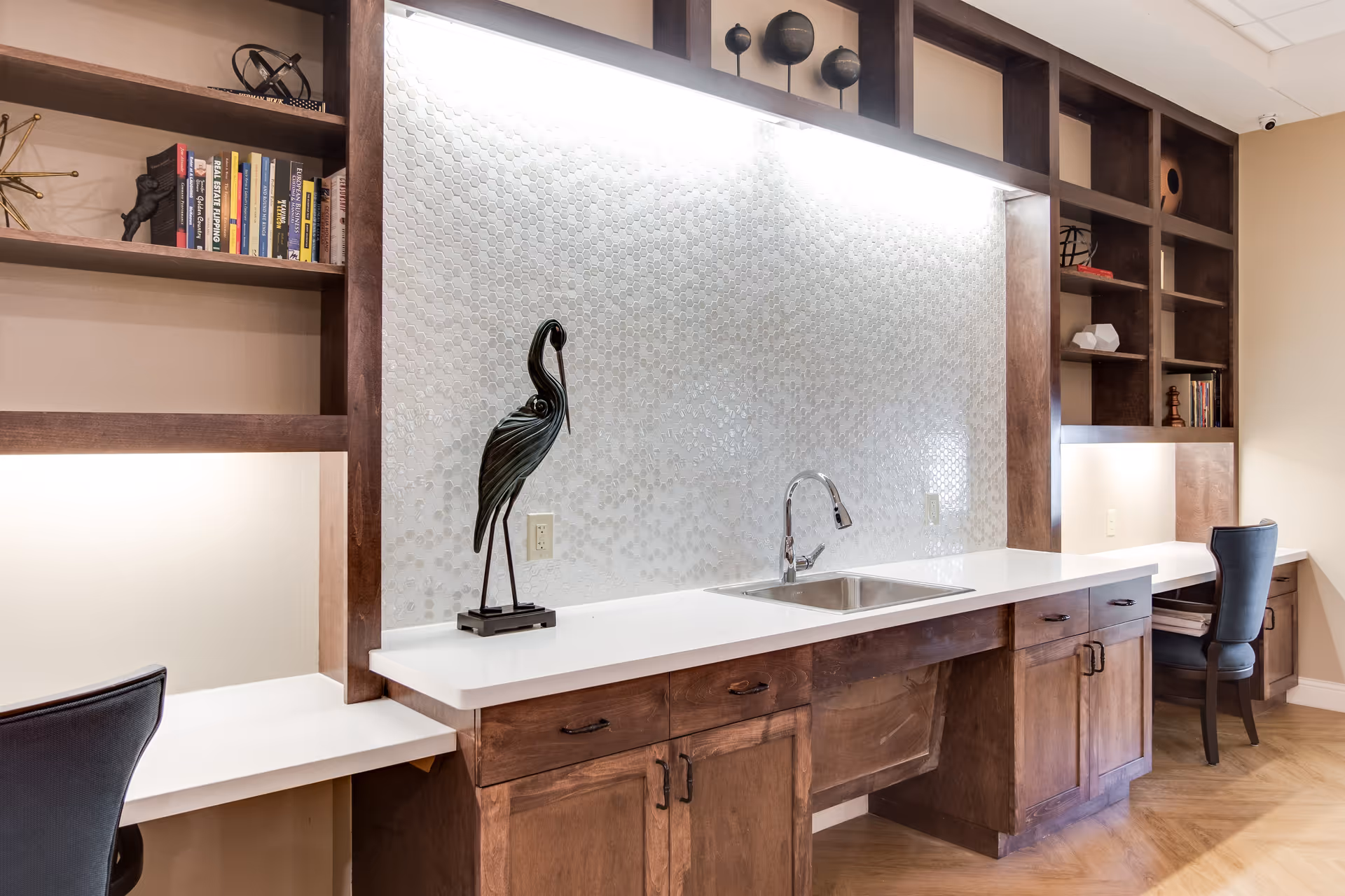 Well-lit interior workstation area with a white countertop and sink, built-in wooden shelving, decorative bird sculpture, and two chairs.
