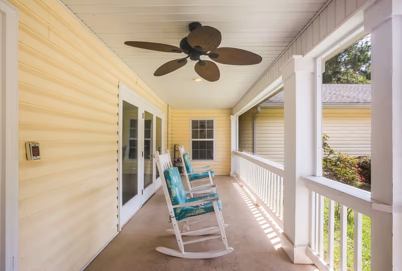 Covered porch with several rocking chairs, a ceiling fan, and white railing overlooking greenery.