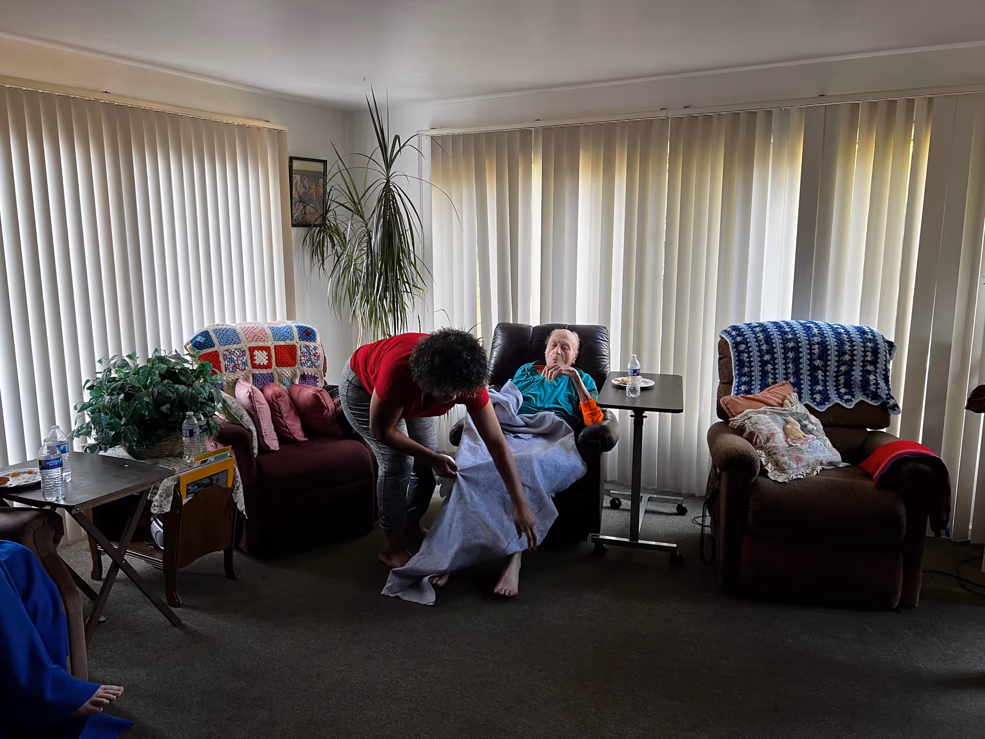 A living room with three armchairs, two covered with crocheted blankets. An elderly person is sitting in the middle armchair covered with a blanket, while a caregiver bends down to adjust the blanket. The room has vertical blinds covering the windows and a small table with a water bottle and a plant nearby.