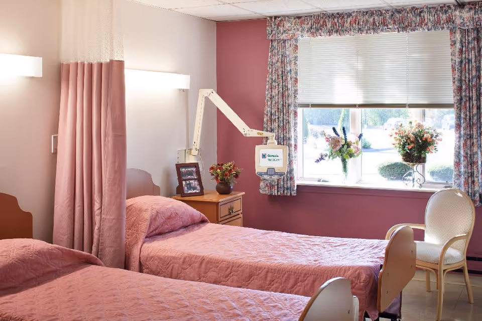 A nursing facility room with two single beds covered in pink quilts separated by a pink privacy curtain. There is a wooden nightstand with a framed photo and a vase of flowers. A medical monitor arm is mounted on the wall. The window has floral curtains and a white blind, with two flower arrangements on the windowsill. A white chair with wooden arms is placed near the window.