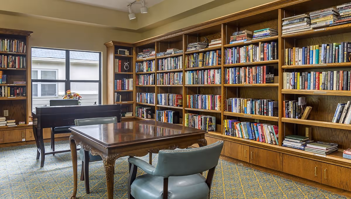 A cozy library room with wooden bookshelves filled with books lining the walls. There is a wooden table with green upholstered chairs around it in the center of the room. A window lets in natural light, and a piano is positioned near the window with a flower arrangement on top.