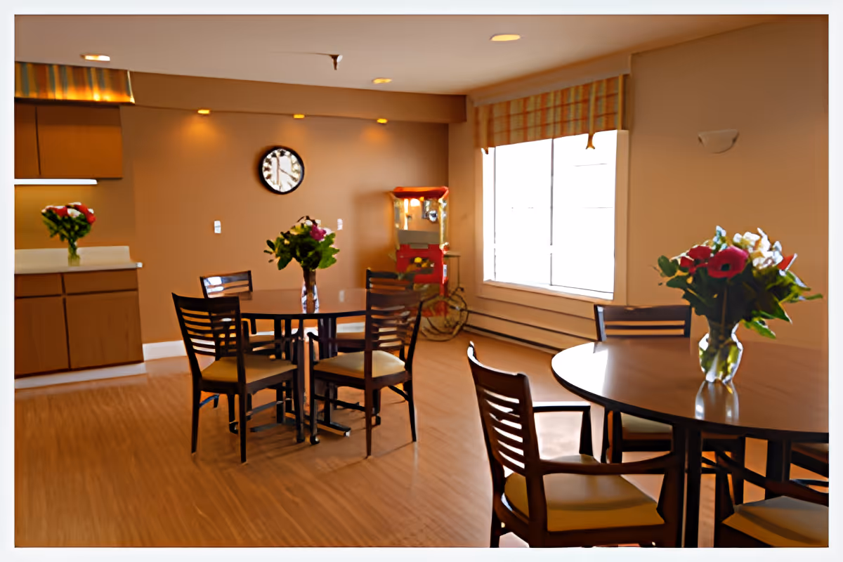 A dining area with two round wooden tables, each surrounded by four chairs. Each table has a vase with colorful flowers. The room has wooden flooring, beige walls, a large window with a striped valance, a wall clock, and a popcorn machine in the corner. There are kitchen cabinets visible on the left side.