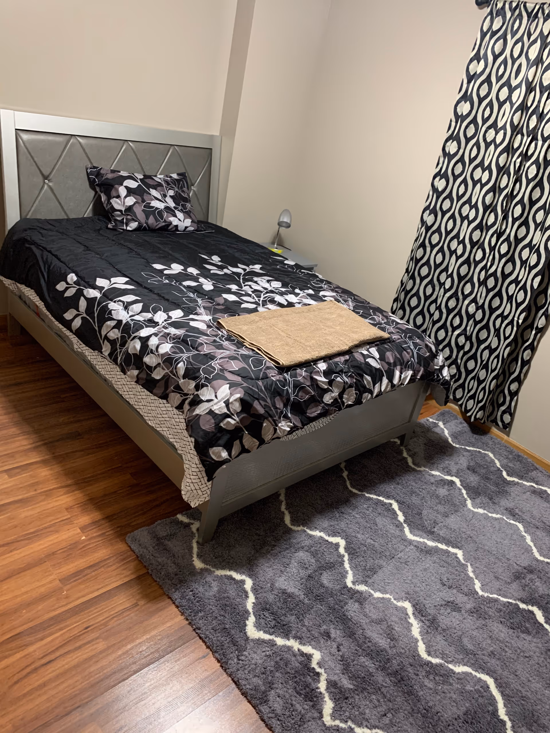 A neatly made single bed with a black and white floral patterned comforter and matching pillow in a small bedroom. A folded beige towel is placed on the bed. The room has wooden flooring, a gray and white patterned rug, a small bedside table with a lamp, and a window with black and white patterned curtains.