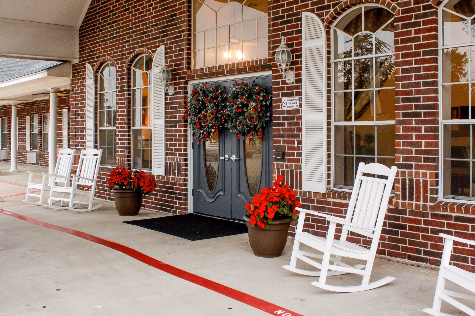 Entrance of Southern Knights Senior Living Community with a brick exterior, double doors decorated with a festive wreath, white rocking chairs on the porch, and potted red flowers on either side of the doors.