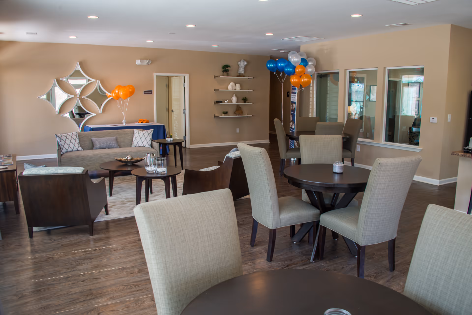 Common area with round tables and upholstered chairs, a sofa seating area, decorative wall mirrors, and orange and blue balloons.