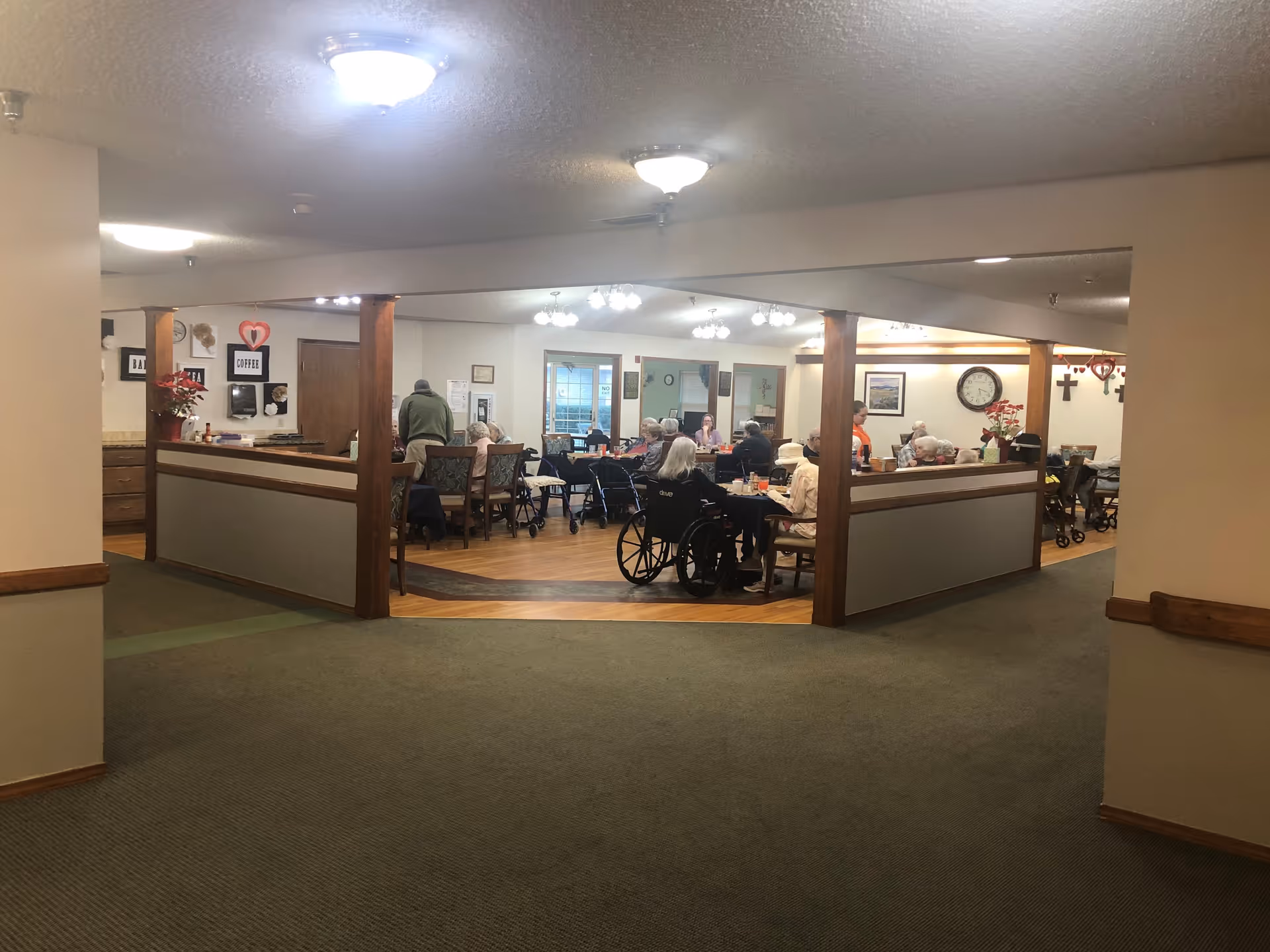 A dining area in a senior living facility with several elderly residents seated at tables eating and socializing. The room has wooden pillars, wall decorations including a clock and framed pictures, and overhead lighting. Some residents are in wheelchairs, and a staff member is assisting them.