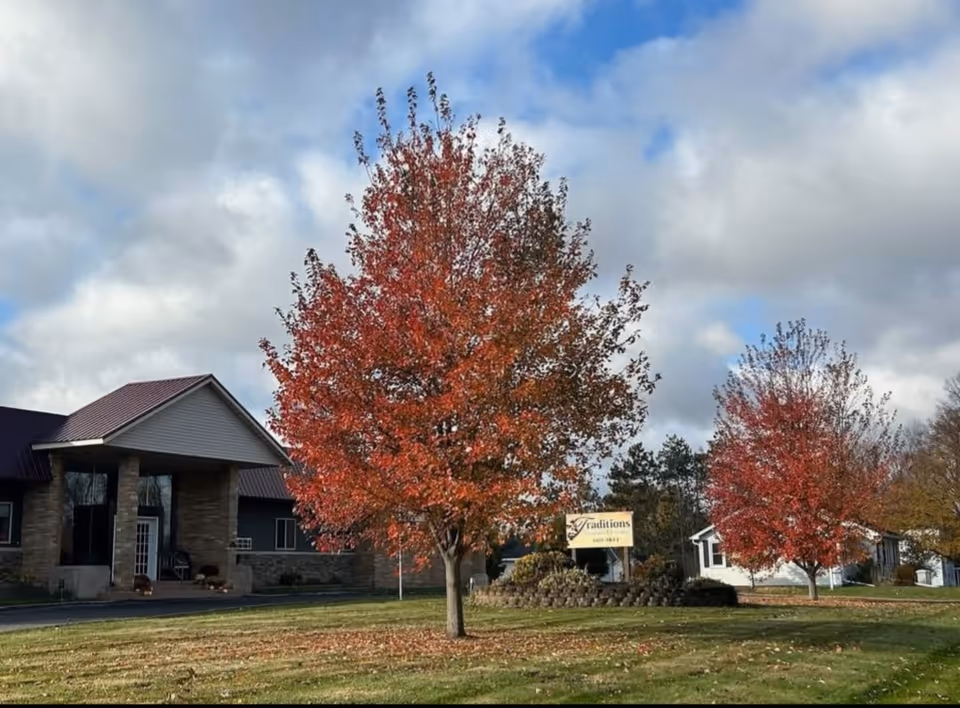 Front exterior of Traditions Assisted Living with red autumn trees, a lawn, and the facility sign under a cloudy sky.