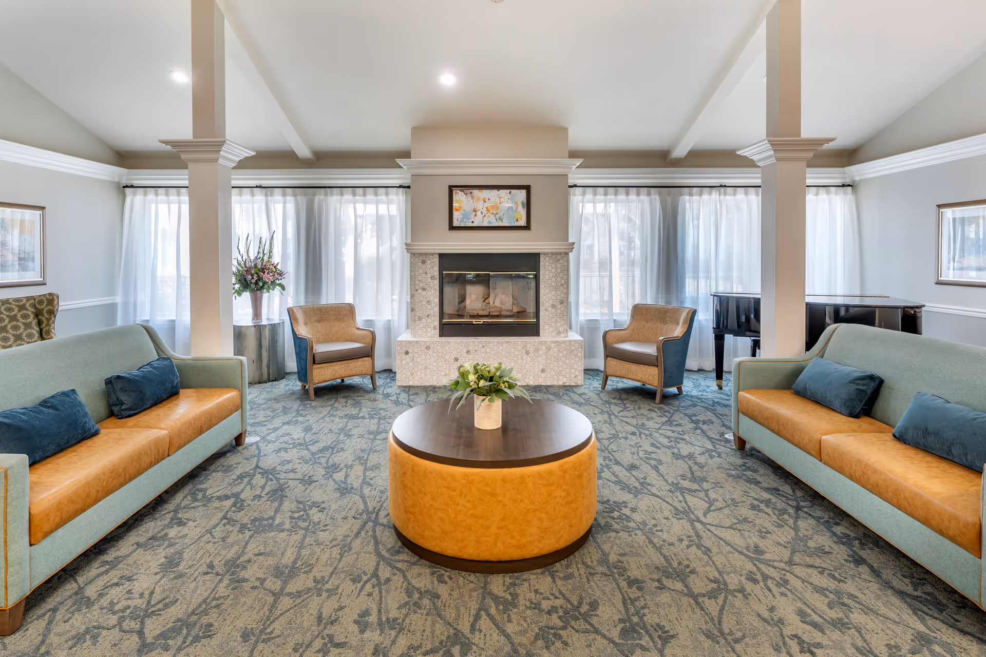 A bright and spacious living room with two light blue and tan sofas facing each other, two beige armchairs near a fireplace with a framed painting above it, a round coffee table with a plant in the center, large windows with sheer white curtains, and a piano in the corner.