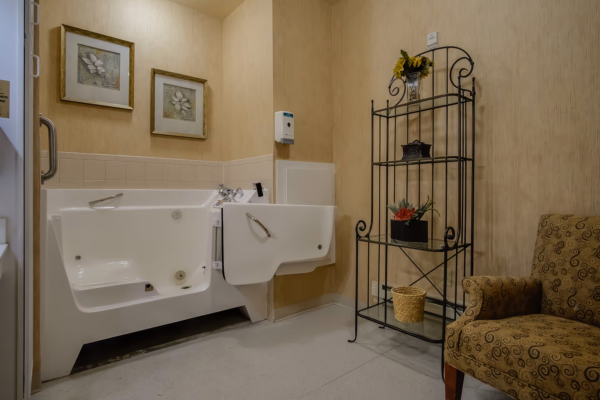 A bathroom with a walk-in bathtub featuring a door for easy access, two framed floral pictures on the wall, a wall-mounted soap dispenser, a decorative black metal shelving unit with plants and a basket, and a patterned upholstered armchair.