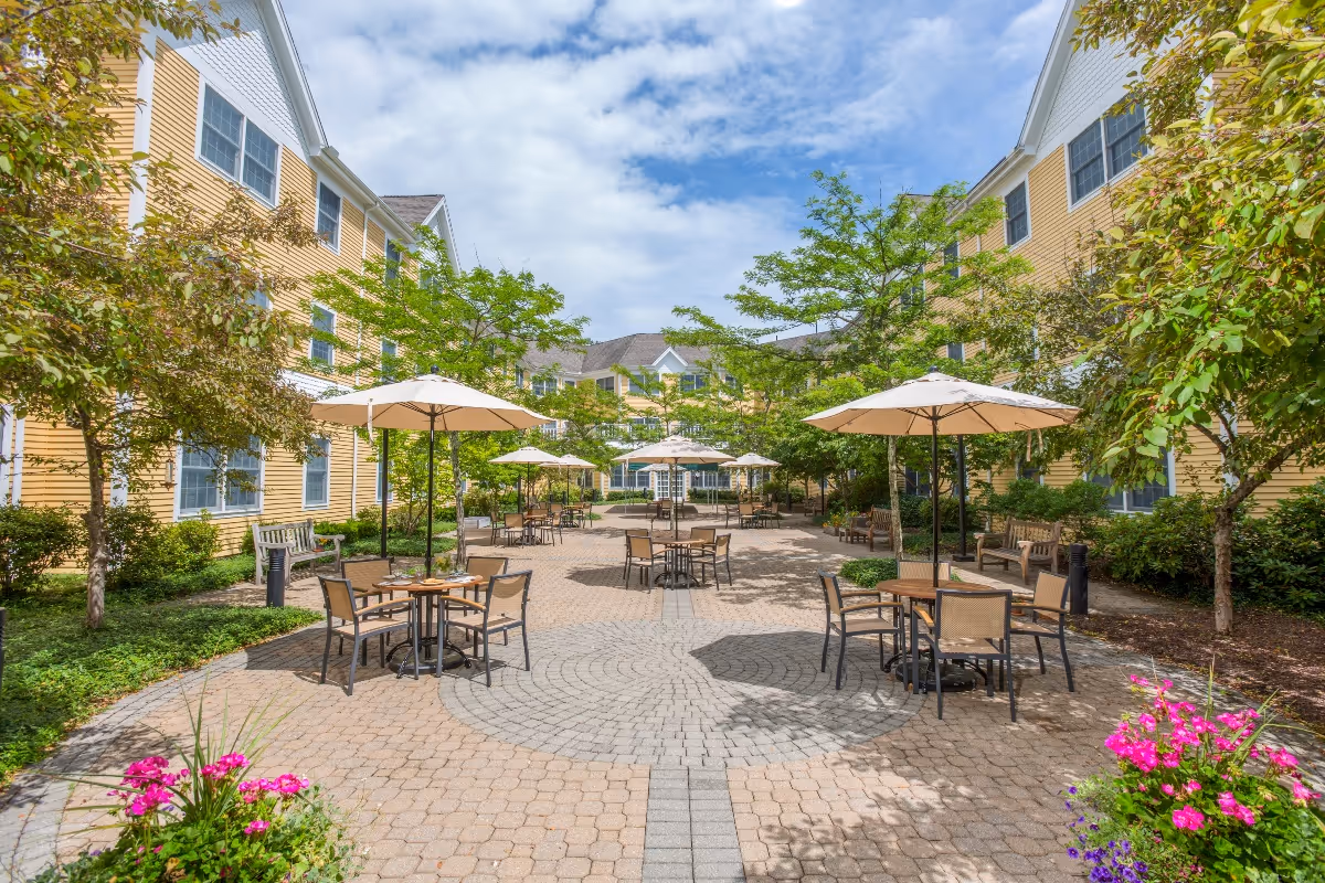 Outdoor courtyard area at River Ridge at Avon with multiple tables and chairs under large beige umbrellas, surrounded by yellow residential buildings, green trees, and colorful flower beds under a partly cloudy sky.