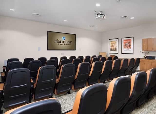A small auditorium or presentation room with multiple rows of black and brown cushioned chairs facing a wall-mounted screen displaying the Harmony Senior Services logo. The room has white walls, a ceiling-mounted projector, two framed posters on the right wall, and wooden cabinets with a countertop and a coffee maker.