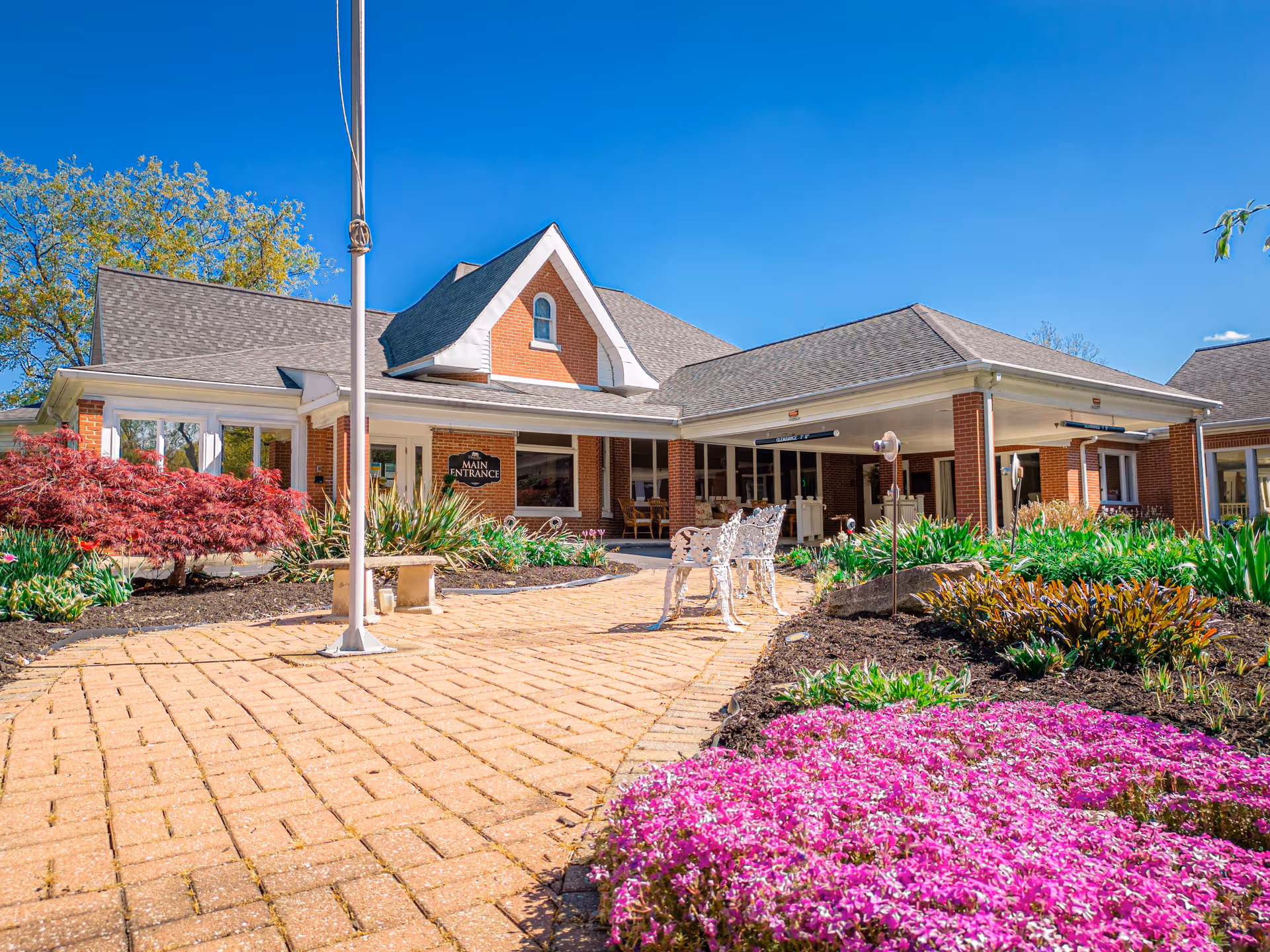 Exterior view of Charles Ford Retirement Communities of New Harmony showing a brick building with a main entrance sign, surrounded by landscaped gardens with colorful flowers and greenery under a clear blue sky.