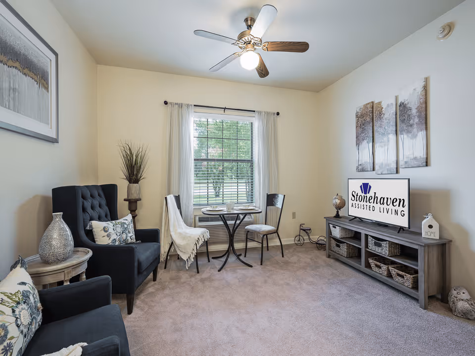 A cozy living room with two dark upholstered armchairs, a small round table with two chairs near a window with white curtains, a ceiling fan with light, and a wooden TV stand holding a television displaying the Stonehaven Assisted Living logo. The room has beige walls, carpeted floor, and decorative items including framed artwork, a vase, and baskets on the TV stand.