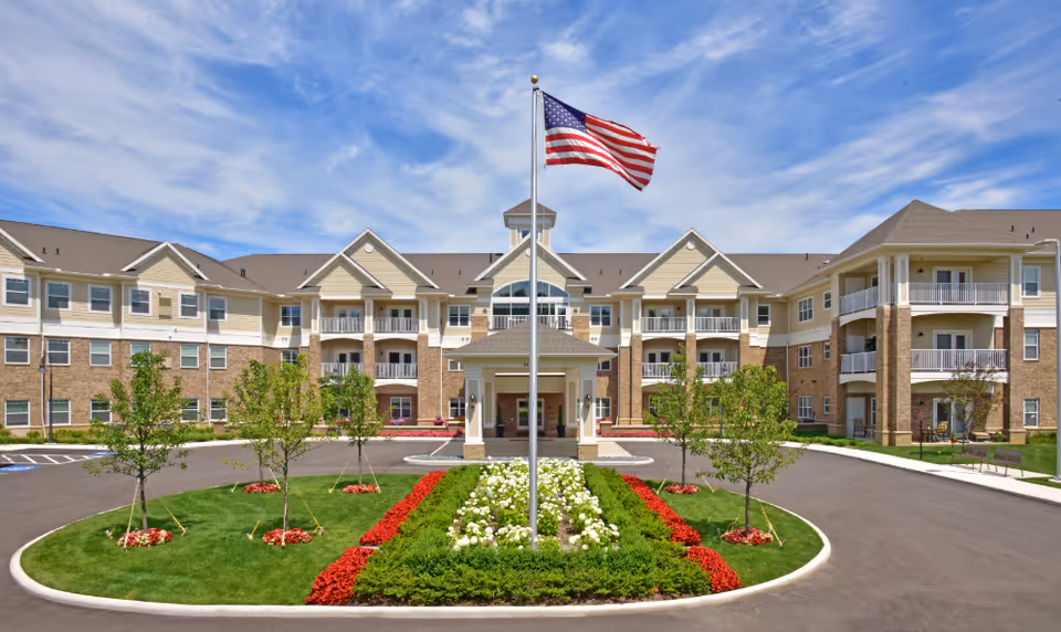 Front exterior view of Rose Senior Living Avon building with a circular driveway, landscaped garden with red and white flowers, young trees, and an American flag on a flagpole in the center under a partly cloudy blue sky.