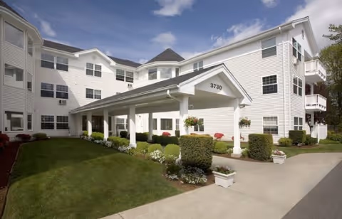 Exterior view of a white multi-story senior living facility with a covered entrance labeled 3730, surrounded by well-maintained green lawns, bushes, and flower pots under a partly cloudy sky.