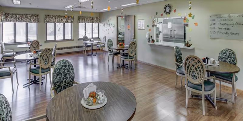 Light-filled dining room with round tables and patterned chairs, hardwood floor, windows and a serving counter.