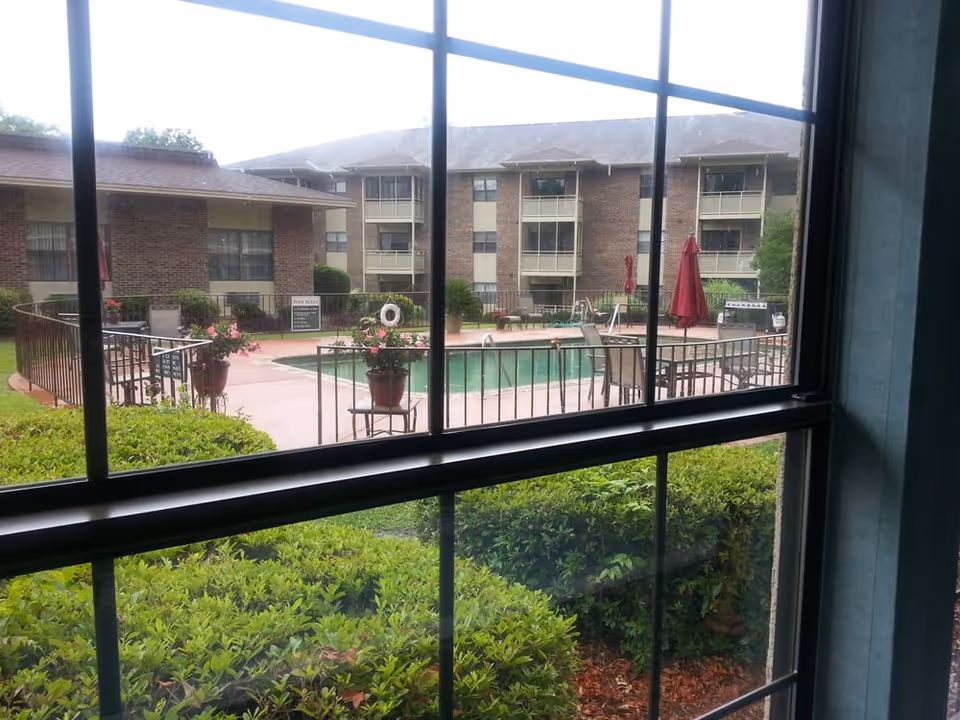View through a window showing an outdoor swimming pool area with patio chairs, tables, umbrellas, and potted plants. Surrounding the pool are brick residential buildings and green bushes in the foreground.