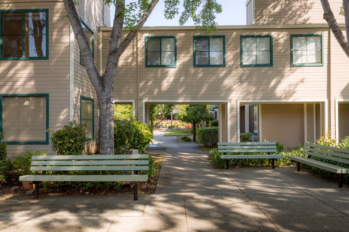 Outdoor courtyard area of a senior living facility with three green benches arranged around a paved walkway. The building has beige siding with green window frames, and there are trees and bushes providing shade and greenery.