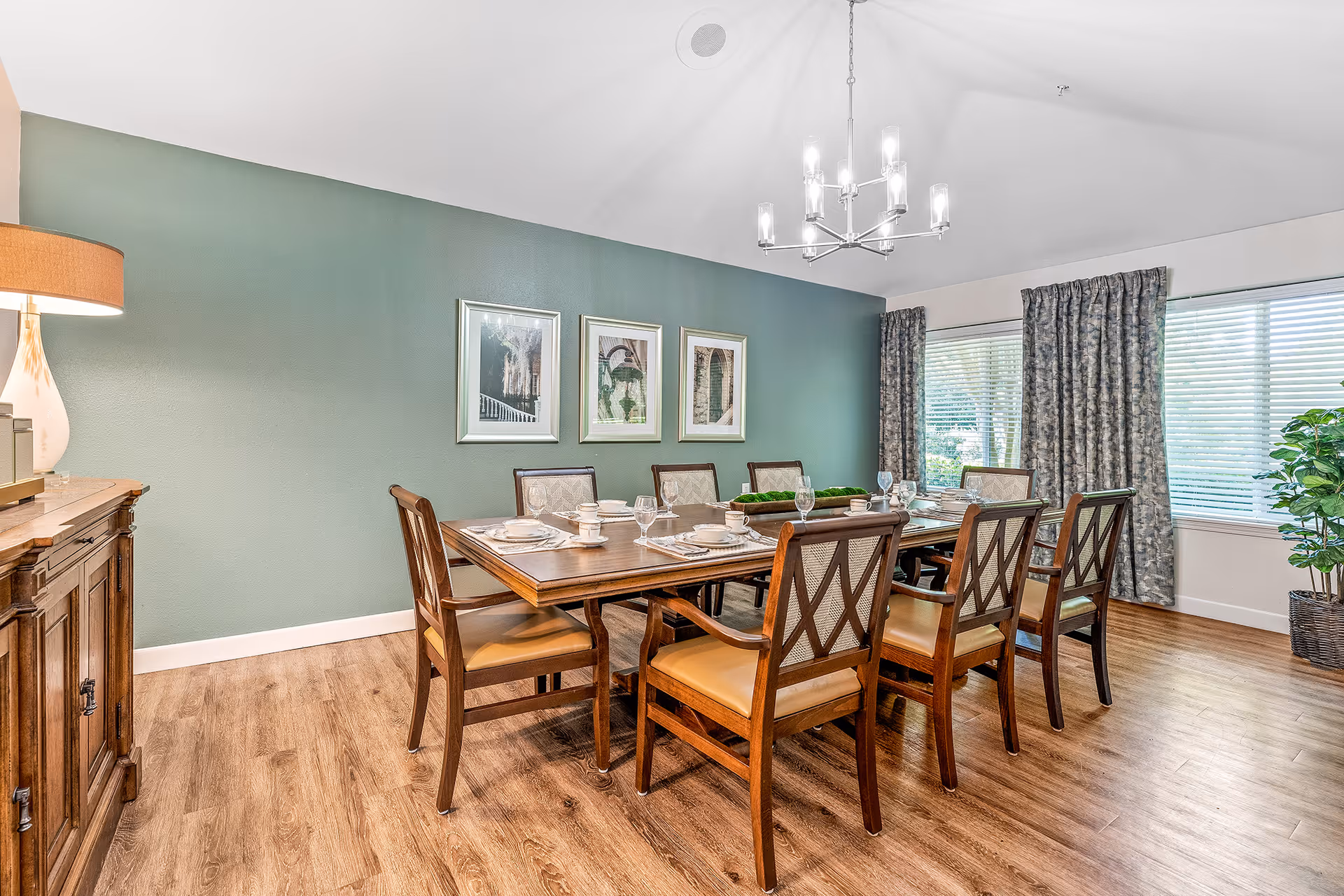 A well-lit dining room with a wooden dining table set for eight people. The table is surrounded by wooden chairs with cushioned seats. On the table are plates, cups, and glasses arranged neatly. The room has a green accent wall with three framed pictures, a wooden sideboard with a lamp on the left, large windows with patterned curtains on the right, and a modern chandelier hanging from the ceiling. The floor is a light wood finish.