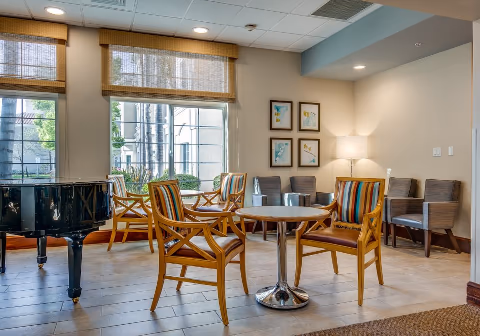 Bright senior living common lounge with a round table, striped wooden chairs, a grand piano, and seating by large windows.