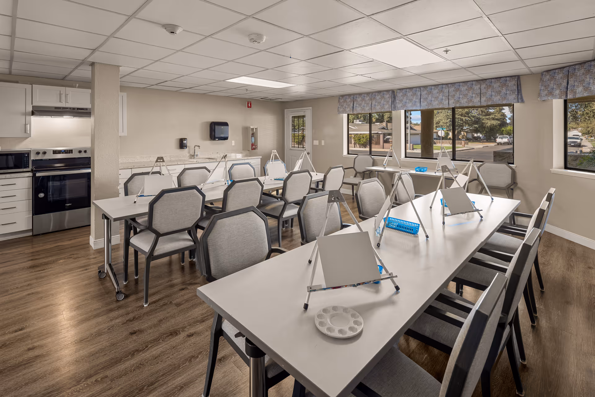 A bright room with several tables and chairs arranged for an art activity, each table has small easels with blank canvases and paint palettes. The room has large windows with a view outside, wood flooring, and a kitchenette area with a stove, microwave, and cabinets.