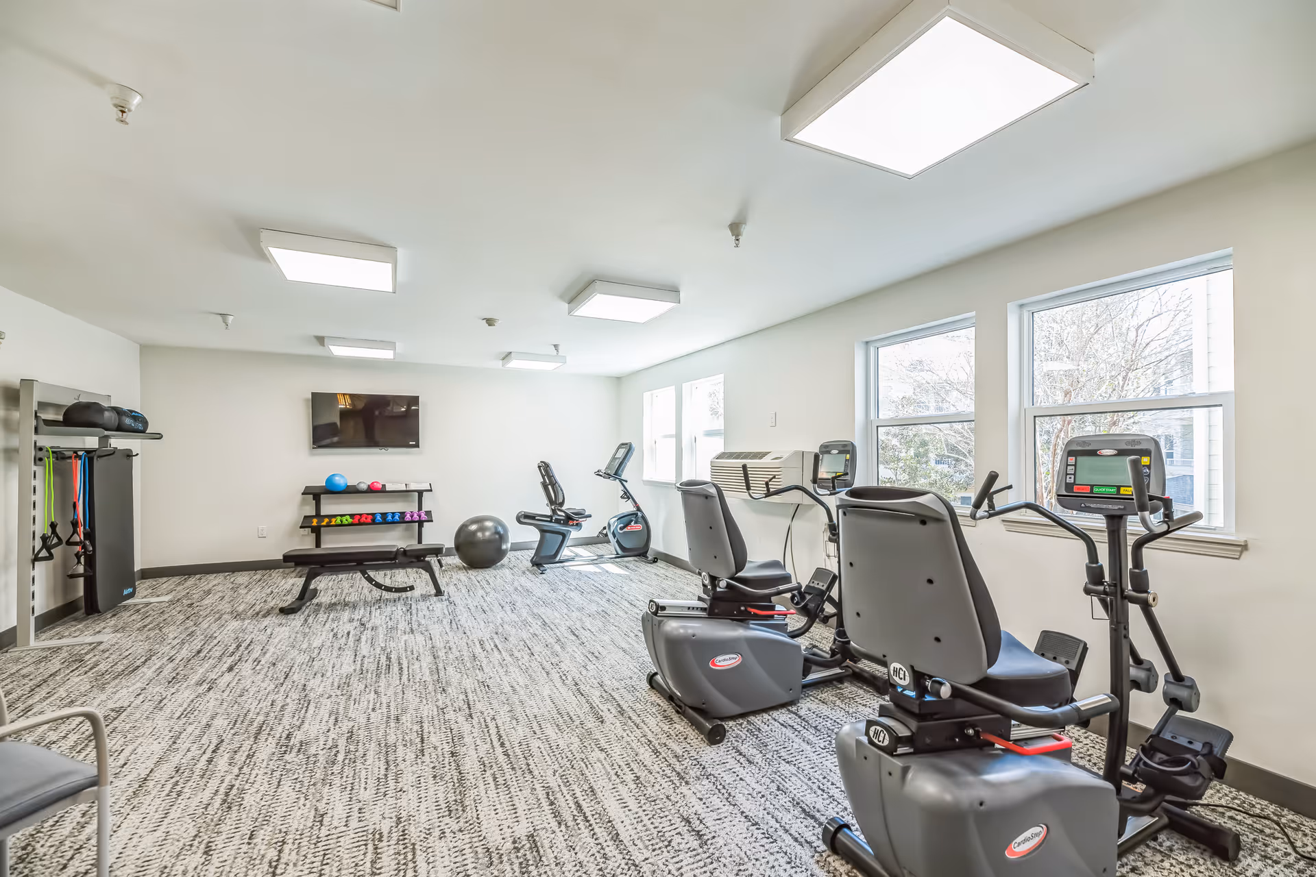 A bright fitness room with exercise equipment including stationary bikes, an elliptical machine, a bench, resistance bands, dumbbells, and exercise balls. The room has large windows letting in natural light and a wall-mounted TV.