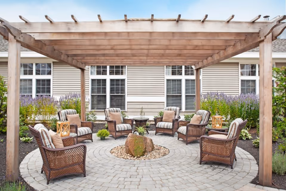 Outdoor seating area with a wooden pergola overhead, surrounded by cushioned wicker chairs arranged in a circle around a central stone feature. The area is paved with circular stone tiles and bordered by plants and flowers, with a building featuring multiple windows in the background.